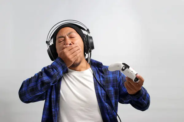 A bored Asian man, wearing headphones, a beanie hat, and a casual shirt, yawns and looks tired while playing video games with a gamepad on a console, feeling disinterested and bored