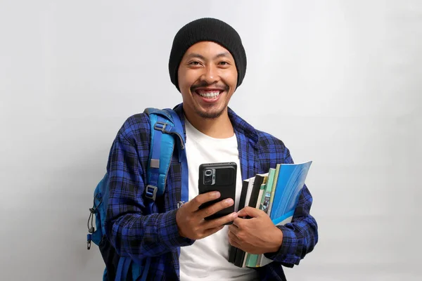 Happy Asian student, dressed in a beanie hat and casual shirt and carrying a backpack, holds textbooks and a mobile phone, smiling at the camera while standing against a white background