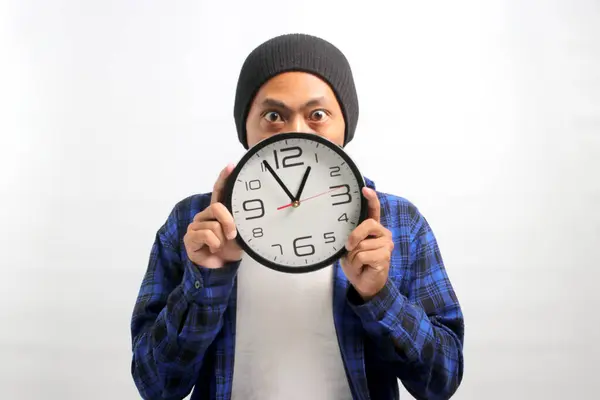 An Asian man, sporting a beanie hat and casual shirt, holds a clock in front of his face, looking at the camera with a surprised and humorous expression, while standing against a white background