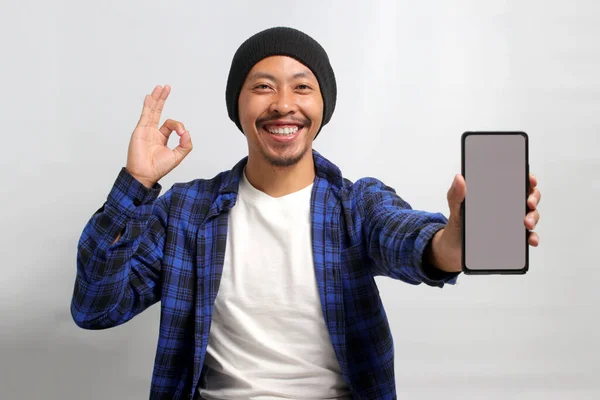 Asian man dressed in a beanie hat and casual shirt displays a smartphone with a blank white screen, creating space for copy or text advertisement, while making an OK sign gesture towards the camera