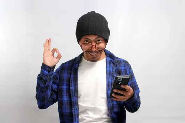 An Asian man, donning a beanie hat, casual shirt, and eyeglasses, flashes an OK sign towards the camera while holding a phone, conveying satisfaction, approval, and a positive review, white background