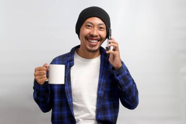 Şapka ve günlük gömlek giymiş Asyalı bir adam, sabah kahvesinin keyfini çıkarırken cep telefonuyla konuşarak birden fazla iş yapıyor, beyaz bir arka planda duruyor.