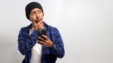 Kafası karışmış ve kafası karışmış Asyalı bir adam, bereli şapka ve günlük gömlek giymiş, cep telefonundaki haberlere tepki veriyor, kafa karışıklığı, mutsuzluk ve derin tefekkür sergiliyor, beyaz arka planda izole edilmiş.