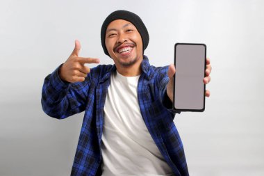 Heyecanlı Asyalı bir adam, bereli bir şapka ve günlük bir gömlek giymiş, boş beyaz ekranlı bir cep telefonuna bakıyor, yeni bir uygulama veya özel bir teklif öneriyor ya da öneriyor.