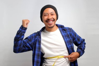 Heyecanlı Asyalı bir adam, bereli bir şapka ve günlük bir gömlek giymiş, karnının etrafında bir mezura tutuyor, beyaz arka planda dururken potansiyel şişmiş midesi hakkında endişelerini dile getiriyor.