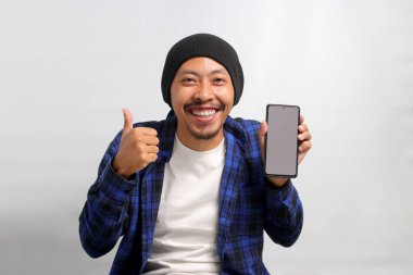 Asyalı heyecanlı genç bir adam, bereli bir şapka ve günlük bir gömlek giymiş, cep telefonunda boş beyaz bir ekran gösteriyor, beyaz bir arka planda dururken kameraya başparmağını kaldırıyor.