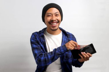 Heyecanlı Asyalı bir adam, bereli bir şapka ve günlük bir gömlek giyer, kameraya gülümserken gururla para dolu bir cüzdanı gösterir, finans, yatırım ve para tasarrufu kavramlarını temsil eder.