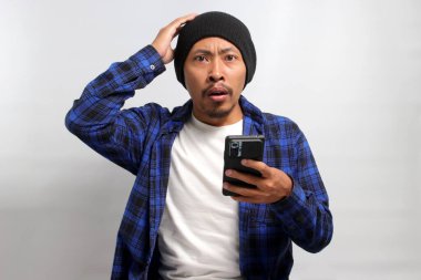 Şapka ve günlük gömlek giyen mutsuz Asyalı adam, akıllı telefonunu tutarken şaşırmış ve üzgün görünüyor, muhtemelen kötü haberlere tepki veriyor ya da online dolandırıcılara kurban gidiyor.