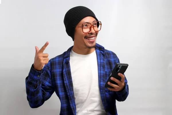 Asian man wearing beanie hat, casual shirt, and eyeglasses points at an empty space while confidently holding his phone, displaying excitement, happiness, and confidence, isolated on white background