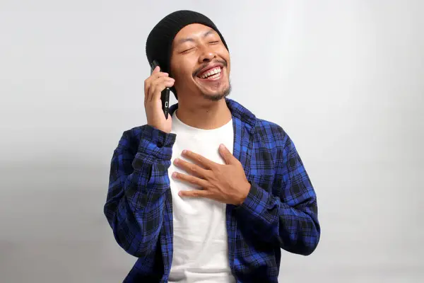 A grateful young Asian man, dressed in a beanie hat and casual shirt, appears relieved, smiling and placing his hand on his chest in response to hearing good news during a phone conversation