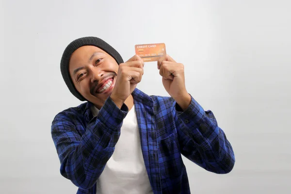 A happy young Asian man, dressed in a beanie hat and casual shirt, enthusiastically shows a credit card in his hand, feeling excited while demonstrating a solution for online shopping payment