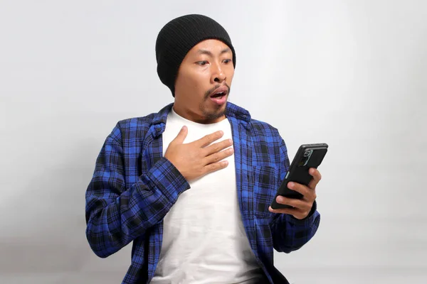 An astonished Asian man, dressed in a beanie hat and casual shirt, reacts with surprise to an ecommerce special offer discount on his phone while standing against a white background