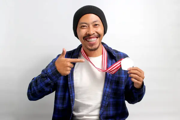 stock image A proud young Asian man, dressed in a beanie hat and casual shirt, smiles as he points at his medal while standing against a white background