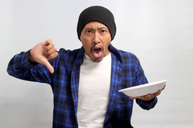 Hoşnut olmayan Asyalı genç bir adam, bereli bir şapka ve gündelik bir gömlek giyer, bir başparmak aşağı vererek memnuniyetsizlik ifade eder, yiyecek için olumsuz bir eleştiri, boş bir beyaz tabak tutarken