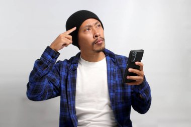 Şapka ve gündelik tişört giyen düşünceli Asyalı bir adam, elinde bir cep telefonu tutuyor, eli çenesinde ciddi ve derin düşünceler taşıyor, mesaj okurken şüphe ve endişe gösteriyor.
