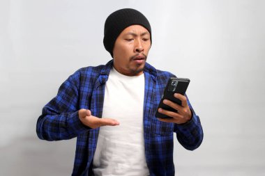 Şapka ve günlük gömlek giyen mutsuz Asyalı adam, akıllı telefonunu tutarken şaşırmış ve üzgün görünüyor, muhtemelen kötü haberlere tepki veriyor ya da online dolandırıcılara kurban gidiyor.