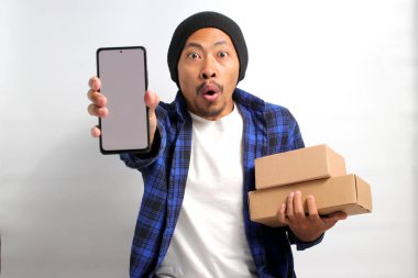 Asyalı heyecanlı genç bir adam, bereli bir şapka ve günlük kıyafetler giymiş, beyaz arka planda duran bir yığın karton kutu tutarken boş beyaz ekranlı bir akıllı telefon sergiliyor.