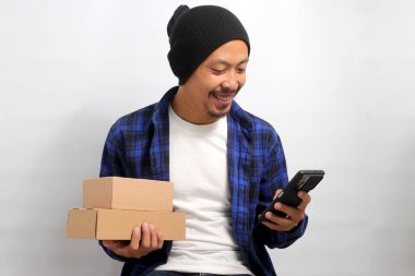 Genç Asyalı bir adam, bereli bir şapka takıyor ve günlük giysiler giyiyor. Birden fazla iş yapıyor. Elinde bir karton kutu tutarken, beyaz bir arka plana karşı telefonunu kullanıyor.