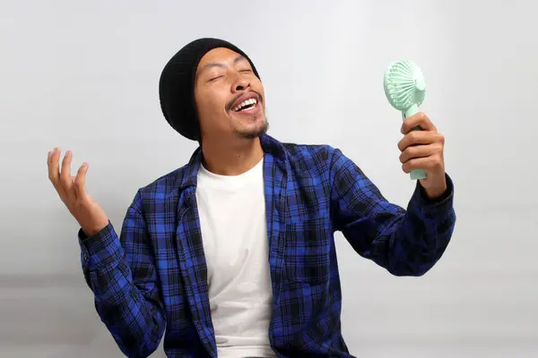 Asian man, dressed in a beanie hat and casual shirt, revels in the airflow and refreshing cool wind from a handheld portable mini fan to beat the summer heat, while standing against a white background