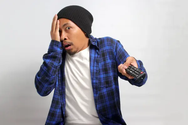 A Scared and frightened young Asian man, dressed in a beanie hat and casual shirt, holds a TV remote while watching a horror movie, peeking through his fingers, isolated on a white background