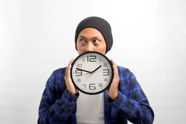 Asian man, dressed in a beanie hat and casual shirt, appears to be deep in thought and planning as he looks aside at an empty copy space, holds a clock in front of his face, partially hiding behind it