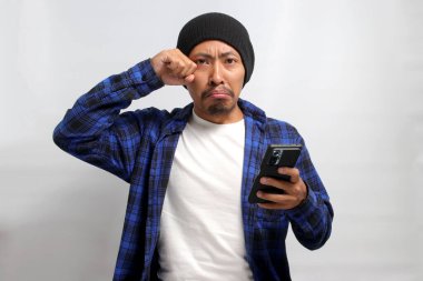 Mutsuz Asyalı bir adam, bereli bir şapka ve günlük bir gömlek giymiş, bir telefon tutuyor, üzgün, endişeli ya da belki de internetten gelen üzücü habere tepki olarak gözyaşı döküyor.