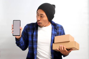 Asyalı heyecanlı genç bir adam, bereli bir şapka ve günlük kıyafetler giymiş, beyaz arka planda duran bir yığın karton kutu tutarken boş beyaz ekranlı bir akıllı telefon sergiliyor.