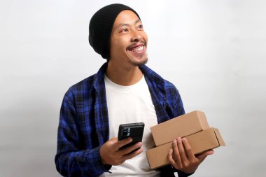 Genç Asyalı bir adam, bereli bir şapka takıyor ve günlük giysiler giyiyor. Birden fazla iş yapıyor. Elinde bir karton kutu tutarken, beyaz bir arka plana karşı telefonunu kullanıyor.