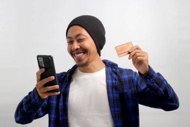 Şapka ve günlük kıyafetler giyen Asyalı genç bir adam, beyaz bir arka plana karşı dururken telefonunu ve kredi kartını online alışveriş için kullanıyor.