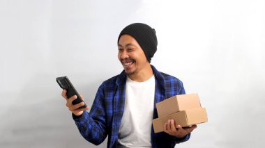 Genç Asyalı bir adam, bereli bir şapka takıyor ve günlük giysiler giyiyor. Birden fazla iş yapıyor. Elinde bir karton kutu tutarken, beyaz bir arka plana karşı telefonunu kullanıyor.