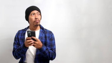 Şapka ve günlük gömlek giyen ilgisiz ve sıkılmış Asyalı bir adam, yanındaki boş alana bakarken cep telefonunu kullanıyor, beyaz bir arkaplana karşı duruyor.