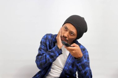 Sıkılmış genç Asyalı bir adam, bereli bir şapka ve günlük bir gömlek giyer, elinde uzaktan kumanda ile televizyon izlerken farklı kanallarda dolaşır, ilginç bir program arar, beyaz arka plan,