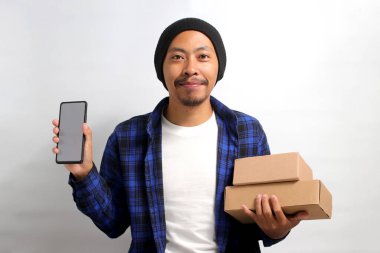 Asyalı heyecanlı genç bir adam, bereli bir şapka ve günlük kıyafetler giymiş, beyaz arka planda duran bir yığın karton kutu tutarken boş beyaz ekranlı bir akıllı telefon sergiliyor.