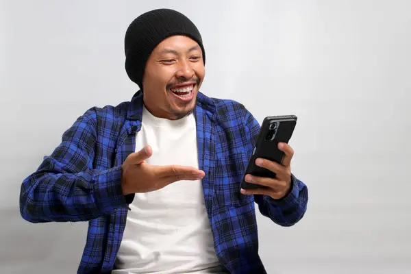 An astonished Asian man, dressed in a beanie hat and casual shirt, reacts with surprise to an ecommerce special offer discount on his phone while standing against a white background