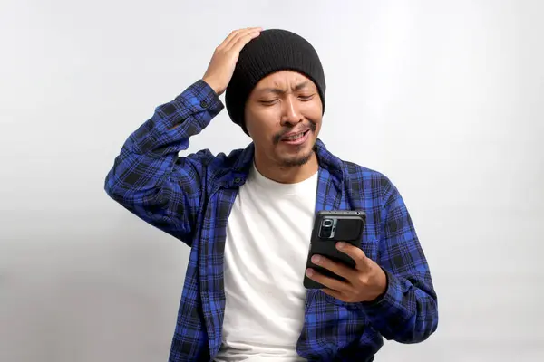 An unhappy Asian man, dressed in a beanie hat and casual shirt, reacts to a message with bad news on his mobile phone, displaying feelings of sadness, worry, and anxiety, Isolated on white background