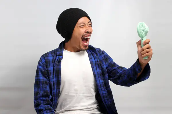 Angry Asian man, wearing a beanie hat and casual shirt, shouts and yells while holding a handheld portable mini fan, frustrated by the intense heat of the summer weather, isolated on white background
