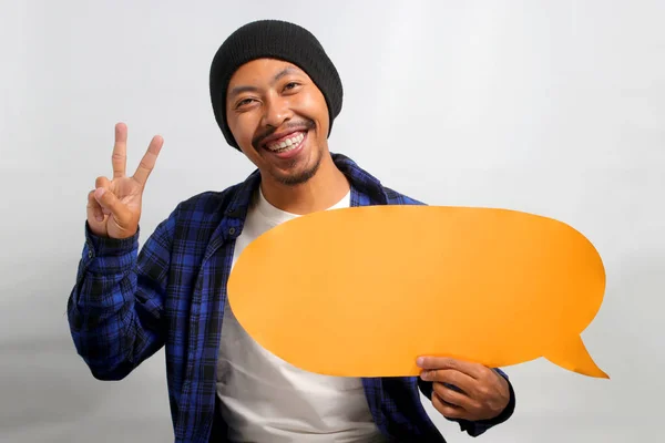 Asian man, dressed in a beanie hat and casual shirt, smiles at the camera and makes a peace V sign gesture while holding a speech bubble, standing against a white background