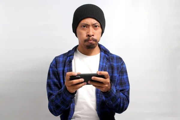 Bored Asian man, wearing a beanie hat and casual shirt, appears bored as he watches an uninteresting video or plays a dull game on his phone while standing against a white background