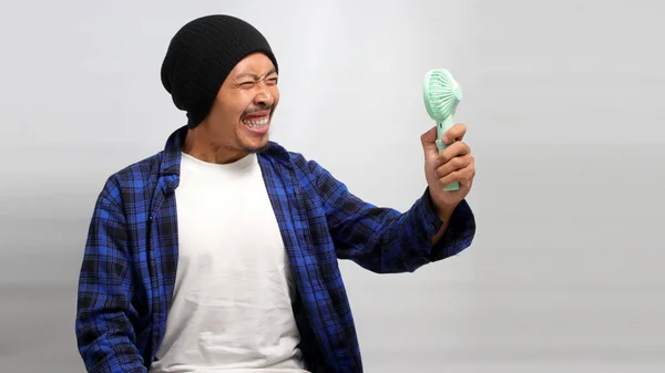 Angry Asian man, wearing a beanie hat and casual shirt, shouts and yells while holding a handheld portable mini fan, frustrated by the intense heat of the summer weather, isolated on white background