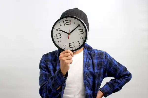 Asian man, dressed in beanie hat and casual shirt, creatively uses a clock to cover his head or face, holding it in front of himself while standing against a white background. Time management concept