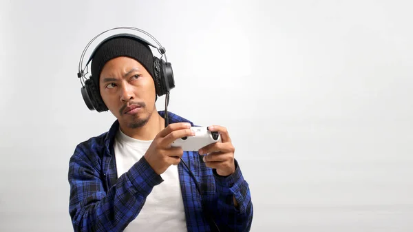 A bored Asian man, dressed in a beanie hat and casual shirt, appears disinterested while playing video games with a gamepad on a console, spending a bored moment, isolated on white background