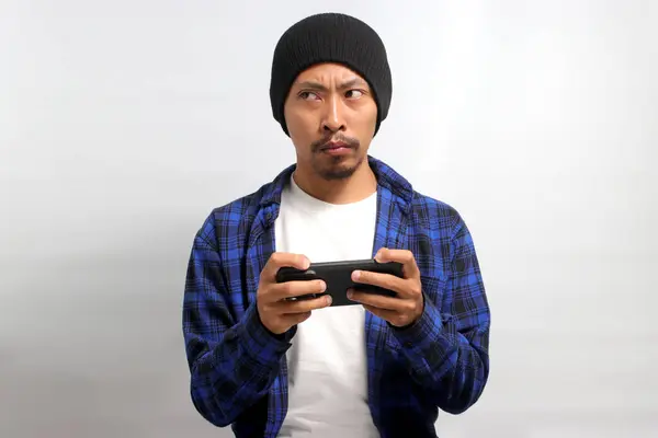 Bored Asian man, wearing a beanie hat and casual shirt, appears bored as he watches an uninteresting video or plays a dull game on his phone while standing against a white background