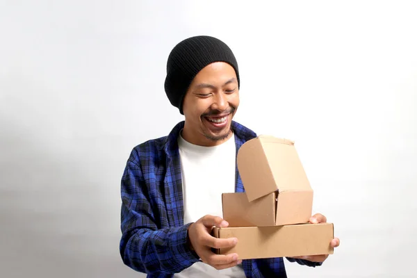 An Asian man, dressed in a beanie hat and casual shirt, smiles while peering into a package parcel cardboard box filled with curiosity. He eagerly unboxes his awaited online store delivery