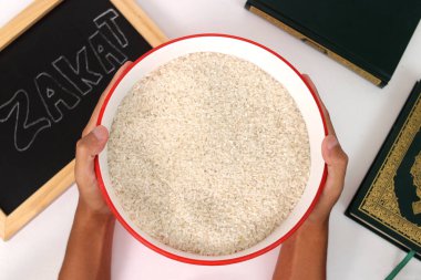 Hand holding a bowl of rice for Zakat al-Fitr, with the Holy Quran and Zakat sign on a white background. Zakat Fitrah is a mandatory form of charity given by Muslims during Ramadan clipart