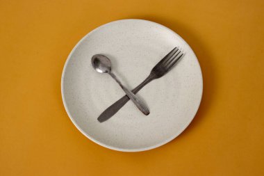 Sarı arka planda kaşıklı ve çatallı boş tabak, Ramazan ayında oruç tutma ve oruç tutma beklentisini temsil ediyor.