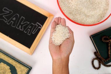 Hand holding a pile of rice for Zakat al-Fitr, with the Holy Quran, prayer beads or tasbih, and Zakat sign on a white background. Zakat Fitrah is a mandatory form of charity during Ramadan clipart