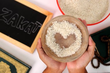 Hand holding a bowl of rice with a heart shape in the center for Zakat al-Fitr, with the Holy Quran and Zakat sign on a white background. Zakat Fitrah is a mandatory form of charity during Ramadan clipart