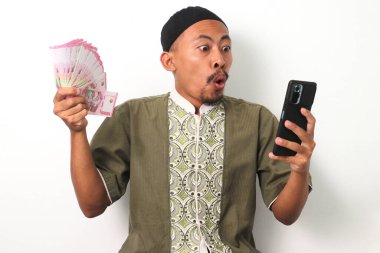Koko tişörtlü ve peci 'li heyecanlı Endonezyalı Müslüman adam bir telefon ve Endonezya rupia banknotları tutuyor ve mali başarıyı kutluyor. Beyaz arkaplanda izole