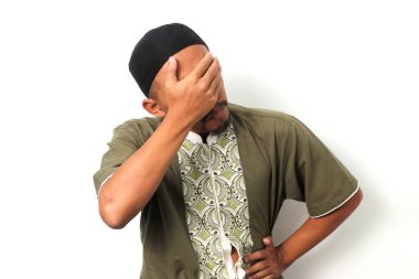 Utanmış Endonezyalı Müslüman bir adam Koko gömleği ve peki ile yüzünü facepalm ile kapatmış. Beyaz arkaplanda izole
