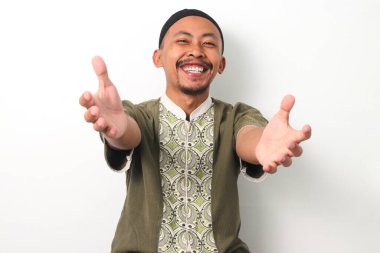 Koko tişörtü ve peki giymiş samimi, güler yüzlü Endonezyalı Müslüman adam kollarını kameraya doğru uzatarak kucaklaşmaya davet ediyor. Beyaz arkaplanda izole